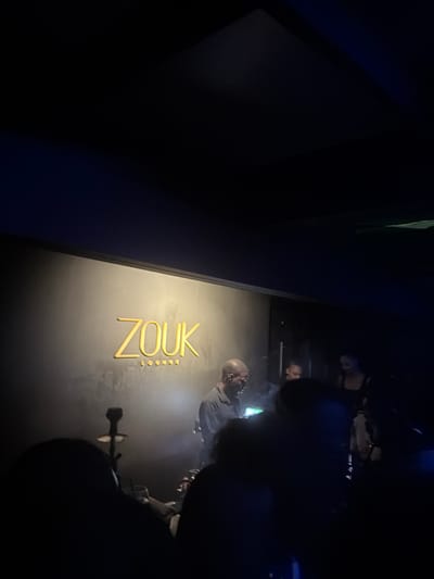 The image shows the "Zouk Lounge" in Holetown, Barbados. A wall displays the lounge's name in gold lettering. Several people are present, engaged in what appears to be social activities. A hookah or similar smoking device is visible near the lower right of the frame. The environment is dimly lit.