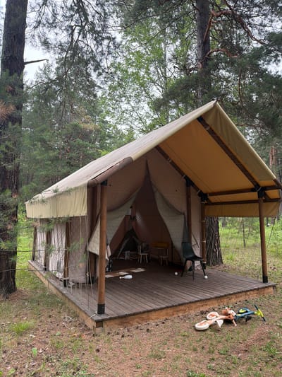 This is a full-shot photograph of a glamping tent set up in a forest. The tent has a wooden platform that extends outwards, with a tan canvas roof and beige walls. Inside the tent, a black chair is visible, along with two small white balls on the wooden floor. Outside the tent, on the grass, lies a dismounted children's balance bike. The environment is characterized by tall pine trees and green foliage, suggesting a natural, outdoor setting. There are no people visible in the image. The scene is located in the city of Atamanovo, Russia.