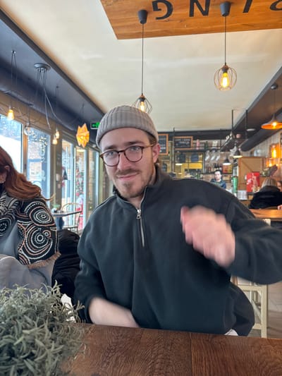 An indoor scene depicts a male subject seated at a wooden table in a cafe or restaurant in the city of Raska Municipality, Serbia. The male, positioned centrally, is smiling and looking directly at the camera. He wears a light grey knit beanie, tortoise-shell eyeglasses, and a dark-colored fleece pullover with a half-zip closure. He has a beard and short brown hair. His right hand is raised and blurred, indicating movement or a gesture. To his immediate left, a portion of another individual, likely female, is visible, displaying reddish-brown hair and a garment patterned with concentric circles. In the foreground, a small potted plant with light green, narrow leaves rests on the wooden table. The background features large windows revealing an outdoor scene with snow and string lights, multiple industrial-style pendant lights with exposed bulbs hanging from the ceiling, and a partially visible bar area with bottles and glassware. Other patrons are indistinctly visible in the deeper background. A wooden sign with partially inverted black lettering is mounted on the ceiling. The environment is well-lit by both natural light from the windows and numerous warm artificial light sources.