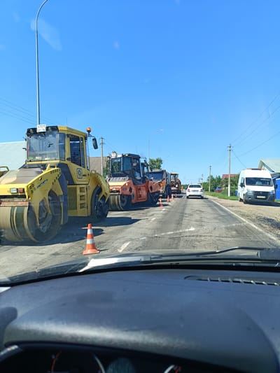 The image depicts a road construction scene on a sunny day in the village of Olyaz, Russia. The foreground shows the interior of a vehicle, suggesting the photo was taken from the driver's perspective. The road surface appears to be undergoing repair, with loose asphalt and tire marks visible. Several heavy construction vehicles are present, including yellow and orange steamrollers and other machinery. Orange traffic cones are strategically placed to redirect traffic. In the background, a white car is driving away from the camera, and a white van is parked on the side of the road. Power lines stretch across the blue sky, and buildings are visible in the distance, indicating a rural or semi-rural setting. The overall impression is one of ongoing road maintenance in a small Russian settlement.