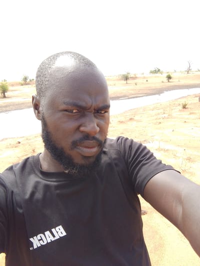 A man is taking a selfie in an outdoor, arid environment. He has dark skin, a shaved head, and a beard. He is wearing a black t-shirt with the word "BLACK" printed on it in white, reversed. The background consists of a landscape with a light-colored, sandy or dirt ground, sparse vegetation, and a shallow body of water or a dry riverbed in the distance. Some small trees are visible on the horizon. Based on the prompt, the location is Mulke Barikin Sayejo, Nigeria.