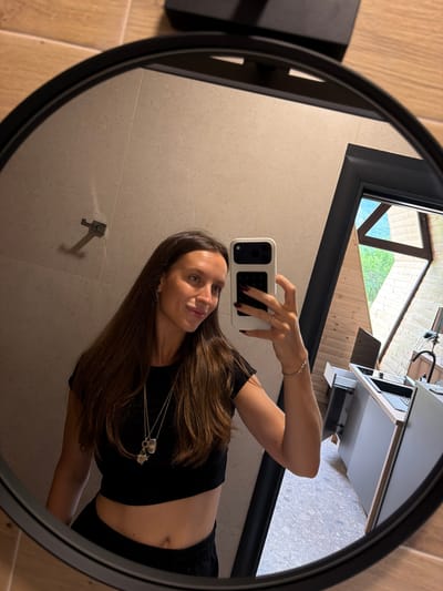 The image, taken in Bistrica, Montenegro, is a reflection in a round mirror. A young woman with long brown hair is visible, holding a smartphone in her right hand to take a selfie. She is wearing a black crop top and black bottoms, with a silver necklace and bracelet. Her left arm is extended, and her ring finger is visible. The background reflected in the mirror shows a modern interior with light-colored tiled walls and wooden floors. To the right, a large angular window reveals a view of lush green trees and a triangular-shaped wooden structure. A kitchen counter with a sink and cabinets is also visible in the reflection.