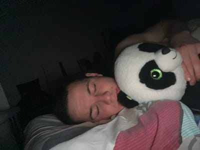 A person is asleep in bed, hugging a plush panda toy. The person is lying on their back, with their eyes closed. The panda toy is positioned against the person's chest. The bed is covered with a striped blanket. The room is dimly lit. It is not possible to confirm the location in Benningen, Germany from the image.