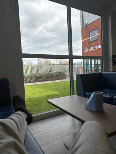 The image, taken from an indoor perspective, looks out through a large window divided by white frames. The view encompasses both interior elements and an outdoor scene. Indoors, the lower half of a person's legs and feet are visible, clad in grey sweatpants and black sneakers, suggesting they are seated. A blue sofa is partially seen, and a light-colored rectangular table occupies the right side of the frame, holding a roll of paper towels.
Outside the window, a prominent red brick building features a large white sign with the word "GREA" and a distinctive red and blue cog-like logo. A dark, rectangular window is also visible on the brick facade. The outdoor landscape includes a vibrant green grassy area, a metallic fence, and a line of dense bushes. Bare trees are discernible in the distance under an overcast but bright sky, indicating daytime. The scene suggests a commercial or industrial setting, possibly a waiting area or office lounge, in Connah's Quay, United Kingdom. No specific activity is ongoing apart from the person relaxing indoors.