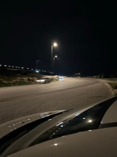 The image displays a night-time scene of a road, presented in a portrait orientation, appearing as if rotated 90 degrees clockwise from a natural landscape view. The left two-thirds of the frame are consumed by a deep black night sky. A prominent, bright circular streetlight, supported by two dark poles, emits a strong white glow in the mid-left portion of the image.

To the right of this primary light source, a paved road or highway occupies the right third of the frame, stretching vertically from the top downwards. The road surface is a textured gray, showing a gentle curve. Along the left edge of this road (when viewed as if correctly oriented), a series of smaller, evenly spaced bright streetlights create a line of white and yellow streaks receding into the distance, possibly indicating motion blur or a longer exposure time.

The far right side of the image is filled by the light-colored bodywork of a vehicle, likely a car, which acts as a large, smooth, reflective foreground element. Numerous small, bright reflections of the distant streetlights are visible across this surface. Faint outlines of green vegetation and indistinct structures can be seen alongside the road further in the background. Two other vehicles, distinguished by their dim headlights and shapes, are faintly visible on the road ahead of the brighter streetlight. The overall impression is a quiet nocturnal journey or a stop on an illuminated roadway. No visible text is present in the image.