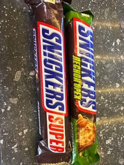 Two Snickers candy bars are positioned vertically on a speckled dark gray surface, possibly a countertop. The left candy bar is mostly obscured by the right one. The visible portion of the left bar has a dark brown wrapper with the "SNICKERS" logo in large blue letters outlined in white and red. Below the logo, the word "SUPER" is printed vertically in red letters. The right candy bar has a green wrapper with the "SNICKERS" logo in similar blue lettering. To the left of the "SNICKERS" logo on the green wrapper, there is text in yellow letters that reads "ПЕCHOH OPEX". Below this text, the wrapper features a close-up image of the candy bar's ingredients, showing nougat, caramel, peanuts, and chocolate. The background is a dark, textured surface with scattered light-colored specks. There are no people or animals present in the image.