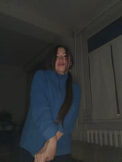 The image shows a young woman with long, dark hair, wearing a blue sweatshirt. She is positioned in a way that her face is tilted upwards and her body is angled towards the viewer. Her arms are crossed in front of her. The background appears to be indoors, with a ceiling that has some structural elements and a blue stripe. There are also some horizontal lines suggesting ventilation or shelving. The lighting is dim, creating shadows and highlighting parts of her face and arms. The overall mood is casual and personal, possibly a selfie. The provided location context is Япрыково, Russia. No text is visible in the image.