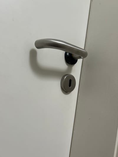 The image shows a close-up, eye-level, outdoor shot of a white door with a brushed metal door handle and a silver keyhole cover. The door handle is curved and appears to be made of stainless steel. It is attached to a black base with visible screws. Below the handle, a circular silver plate covers the keyhole. The keyhole is elongated and oriented vertically. The door is closed. The environment appears to be an urban setting, possibly residential or commercial, in Schöningen, Germany. The lighting is natural, indicating daytime. The image is sharp and well-focused on the door hardware.