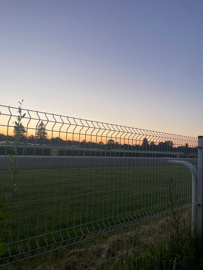 The image depicts a fence in the foreground, with a grassy field beyond. In the background, a treeline is visible against a gradient sky, transitioning from a pale yellow at the horizon to a soft blue at the top. The warm hues of the sunset suggest it is either dawn or dusk. The scene is tranquil, with no visible activity or people. The location is identified as Wysoka, Poland. There is no visible text in the image.