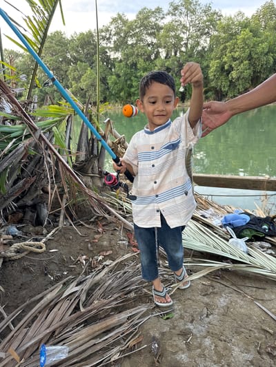 A young boy, approximately 5-7 years old, stands on the muddy bank of a body of water in Grong Grong Capa, Indonesia, proudly holding a small fish he has caught. He is wearing a striped short-sleeved shirt, blue jeans, and flip-flops. A fishing rod with a blue handle and red reel lies beside him. The environment appears to be a natural setting with lush green trees in the background and palm fronds scattered on the ground. The water is calm and reflects the trees. An adult's hand is visible near the boy, perhaps assisting him or congratulating him on his catch. The scene suggests a peaceful and enjoyable fishing excursion.