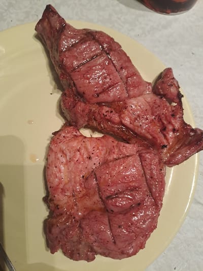 The image shows two pieces of cooked meat served on a light-colored, possibly cream or light yellow, oval-shaped plate. The meat is reddish-pink, indicating it may be cooked medium-rare or medium, or it could be a type of meat that maintains this color after cooking, such as tongue. It features visible grill marks and is generously seasoned with what appears to be ground black pepper or other dark spices. The surface of the meat is glistening, suggesting juiciness or a recent cooking. Some meat juices are visible on the plate beneath the pieces.
The plate rests on a light-colored surface, possibly a tablecloth with a subtle pattern or a plain countertop. A dark, partially visible object, which appears to be a cup or bowl with a patterned surface, is situated to the left of the plate. The scene suggests a meal ready to be eaten in an indoor setting. There is no visible text in the image. The location context provided is Polyanki, Russia.