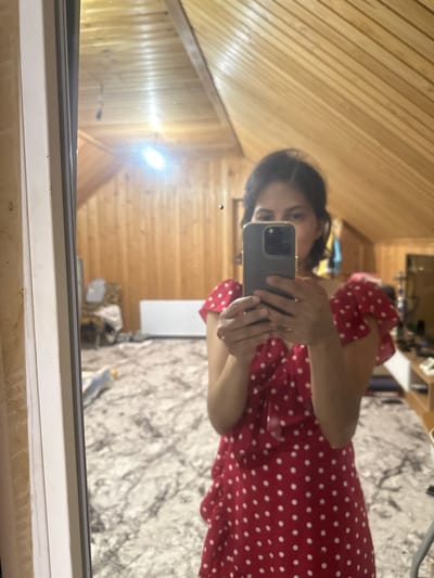 A young woman is taking a mirror selfie in a room with wood-paneled walls and a slanted ceiling. She is wearing a red dress with white polka dots and has short, dark hair. She is holding a dark-colored smartphone in both hands to capture the reflection. The room appears to be a bedroom or living area, with a patterned rug on the floor, a white radiator, and various items of furniture and decor visible in the background. The lighting suggests it might be evening or a dimly lit indoor space, with a light source reflecting brightly on the ceiling.