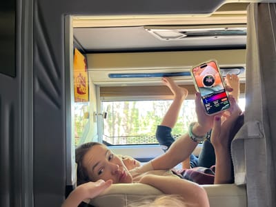 The image shows a woman lying down, looking at her phone. Her feet are propped up in the air, and she appears to be in the back of a vehicle, possibly a bus or RV, given the interior paneling and window view. There is another person, likely a child, lying next to her. The window shows a blurry view of trees and foliage, suggesting an outdoor environment. The phone screen displays what looks like a stock trading app or cryptocurrency tracker. The woman is wearing bracelets on her wrist. The lighting suggests it is daytime. There are no visible landmarks or specific identifiers to place the location within Mùi 3, Vietnam, or any other city.