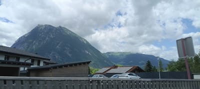 The image depicts a scenic view in Golling an der Salzach, Austria, featuring a prominent mountain in the background, partially obscured by clouds. In the foreground, there are several buildings, including a white house with a dark roof and a long, wooden-clad structure. A white Tesla is parked near a house with solar panels on its roof. A wooden fence runs horizontally across the bottom of the image, suggesting the viewpoint is from a road or elevated area. A traffic sign with a blank face is visible on the right side of the frame. The sky is overcast with patches of blue. The environment is characterized by a mix of natural mountain landscape and residential structures.