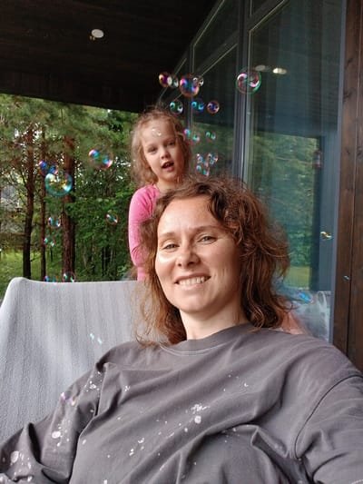 The image shows a woman in the foreground, seated on a gray blanket-covered chair. She is wearing a dark gray t-shirt splattered with white paint. Behind her, a young girl in a pink shirt stands, looking towards the camera with her mouth open. Both are outdoors, possibly on a porch or deck, with trees and greenery visible in the background. Numerous colorful soap bubbles are floating in the air around them. The setting appears to be a residential area, consistent with the town of Verkhneye Dubrovo, Russia.