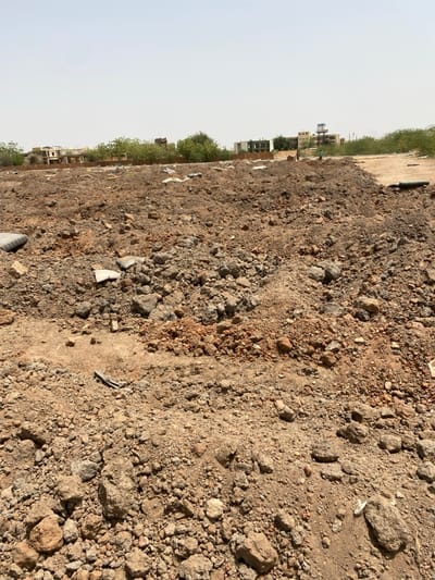 The image shows a large, mostly barren area of land in Khartoum, Sudan. The land is composed of loose, brown dirt and small rocks. There are some scattered pieces of debris visible. In the background, there are several buildings and some trees. Two people are visible in the background, walking in the distance. The sky is clear and blue. The image was taken at coordinates (latitude: 15.528, longitude: 32.571) in Khartoum, Sudan.  The image is likely taken from a slightly elevated vantage point.