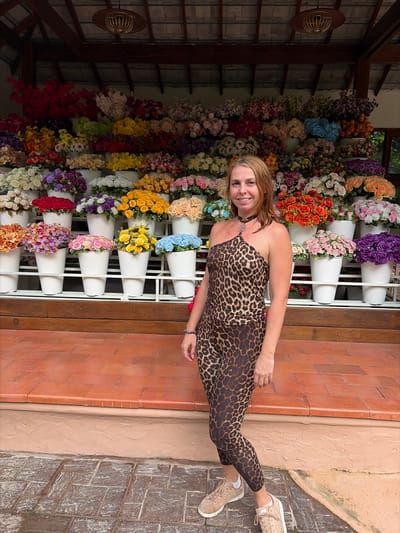 A fair-skinned woman with blonde hair stands in front of a large display of artificial flowers. She is wearing a one-shoulder leopard print top and matching leggings, with beige sneakers. Her necklace is a black choker with a silver pendant. The flowers are arranged in white pots on tiered shelves, creating a colorful backdrop. The setting appears to be an outdoor stall or shop, possibly in Mae Rim, Thailand, given the architecture and abundant greenery visible in the background. The ground in front of the stall is paved with terracotta tiles and cobblestones.