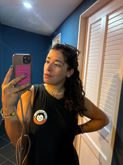A young woman with curly dark hair and light skin is taking a selfie in what appears to be a hotel hallway in Kampot, Cambodia. She is wearing a black tank top with a circular logo of a monkey that reads "MAD MONKEY." She has several bracelets on her left wrist, one of which is made of colorful beads. She is also wearing a necklace. Her right hand is holding a smartphone, which has a pink pop socket on the back. The hallway has dark blue walls and a white door with louvered panels. A bright light is visible on the ceiling.