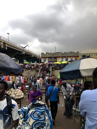 This is a medium shot, taken from a low angle, of a crowded outdoor market in Ojokoro, Nigeria, on a cloudy day. In the foreground, people are walking in various directions, some carrying bags and packages. A man in a blue and white checkered shirt is bending down to pick up a black plastic bag. To his left, a person in a blue shirt with a white design on it is walking away from the camera. In the middle ground, more people are visible, some browsing stalls under colorful umbrellas, and others walking up a set of stairs. The stalls display various goods, including clothing and what appear to be sacks of produce. In the background, a large concrete overpass stretches across the scene, with a few streetlights visible. A building with yellow siding and multiple windows is visible on the right side of the frame. The ground is dirt and appears somewhat uneven.
