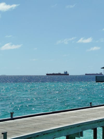 The image shows a serene seascape of the Maldives, with a clear blue sky above dotted with a few wispy clouds. The turquoise waters of the Indian Ocean shimmer under the sunlight, with a gentle ripple across its surface. In the distance, several large cargo ships are visible on the horizon, hinting at the maritime activity in the region. In the foreground, a wooden boardwalk or pier extends into the water, its weathered planks creating a sense of depth and perspective. The overall scene is peaceful and evokes a sense of tranquility, characteristic of a tropical island paradise. The location is Gulhi Falhu, Maldives.