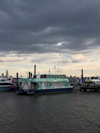 This is a wide shot of a harbor on a cloudy day, with the New York City skyline visible in the distance. In the foreground, a ferryboat is docked. The boat is decorated with an advertisement for Dove products, and the slogan "The Game Is Ours" is visible on its side. The water is dark and choppy, and there are several other boats in the background, including a larger passenger ferry. The sky is filled with dramatic, dark clouds, which are beginning to break apart in some areas, allowing hints of lighter sky to show through. The scene appears to be set in Weehawken, New Jersey, given the proximity to the Manhattan skyline.