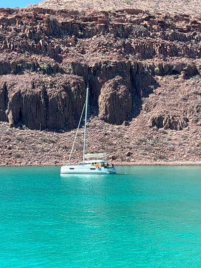 The scene depicts a sailboat anchored close to a rocky shoreline, likely in a cove or inlet, alongside turquoise-colored water. The boat is white with a single mast and rigging, oriented parallel to the shore. Adjacent to the boat is a steep, rocky cliff face consisting of dark-colored, arid-looking rock. The location is Unknown, Mexico. No people are clearly visible.