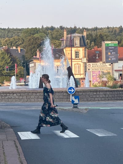 A young woman with brown hair pulled back in a ponytail, wearing a dark floral print dress with off-the-shoulder sleeves and black boots, is crossing a pedestrian street in Baccarat, France. She is walking from left to right, with her body angled towards the viewer. She is holding a colorful object in her right hand.
The scene appears to be a public square or intersection. In the background, there are two multi-tiered fountains spraying water high into the air. Behind the fountains, there are buildings with distinctive architecture, including a building with a mansard roof and cream-colored walls. A clock is visible on the facade of one building. There are also trees and a hillside covered in dense green foliage.
Signage is visible in the background, including a weather display showing "Météo" with temperatures of 36°C for "Matin" and "Après-midi." Another sign indicates directions to "RAON L'ETAPE BERTRICHAMPS" with an arrow to the right and "AUTRES DIRECTIONS" with an arrow to the left. A blue circular sign with a white arrow pointing right is also present. A traffic light showing a red signal is visible on the left side of the image.
The foreground shows a pedestrian crosswalk with white stripes painted on the asphalt. The ground is wet, suggesting recent rain or spray from the fountains. The overall lighting suggests it is daytime, possibly late afternoon.
