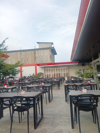 The image depicts an outdoor seating area at a Pertamina gas station in Geulumpangsulu Timu, Indonesia. Numerous tables and black plastic chairs are arranged on a concrete patio, suggesting a dining or rest stop facility associated with the gas station. In the background, a long, single-story building with a red and white facade is visible, bearing the Pertamina logo. Beyond this, a taller, weathered concrete building stands, possibly under construction or renovation. A row of motorcycles is parked to the left of the seating area, along with a white car. Green trees and potted plants add some natural elements to the scene. The sky is overcast, with a few wisps of clouds. The overall impression is that of a functional commercial space.