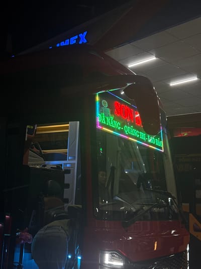 The image shows a close-up of an arcade game, likely a driving simulator, at night. The game features a red truck cab with a brightly lit LED display that reads "XIN CHAO" in Vietnamese, meaning "Hello". The display is bordered by a colorful LED strip. Inside the cab, a person's reflection is visible in the windshield, along with parts of the interior. To the left of the game, the word "EMEX" is displayed in blue LED lights. The scene appears to be indoors, possibly in an arcade or entertainment center, given the lighting and the nature of the game. The overall atmosphere is vibrant and illuminated by the game's lights.