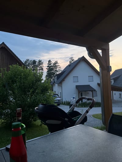 The image shows a view from under a wooden pergola, looking out at a suburban street scene in Milná, Czechia, at dusk. In the foreground, a dark grey table occupies the bottom of the frame, with the tops of two bottles, one red and one green, visible on its left edge. A black baby stroller with a green fabric interior is positioned in the middle ground, to the right of a lush green bush. Beyond the stroller and bush, a modern white house with a dark roof and closed white shutters on its windows is visible. A silver car is parked on the driveway in front of the house. A person in a light blue shirt and white hat is walking on the sidewalk in front of the house. Pine trees form a backdrop behind the houses, and the sky above is a pale blue with scattered clouds, illuminated by the setting sun. The overall atmosphere is peaceful and domestic.