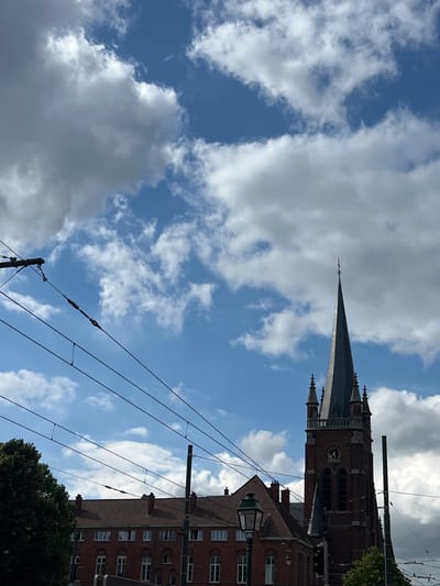 The photograph captures a church tower with a spire, partially obscured by overhead wires, against a sky filled with dramatic clouds. The church appears to be constructed of dark red brick, with visible architectural details such as arched windows and smaller spires or turrets on its upper levels. A clock face is visible on the tower. In the foreground, a multi-story building with a red tiled roof and numerous windows lines the street. A streetlamp with a green lantern is also present. The scene is framed by a dense green tree on the left and utility poles and wires that crisscross the sky, suggesting an urban environment. The specific location is identified as Jette, Belgium, implying the church is likely a local landmark within the municipality. The image does not contain any people or discernible actions.