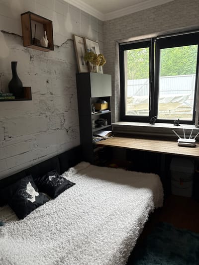 The image shows a bedroom with a bed, a desk, and a window. The bed is covered with a white, fluffy blanket and has two black pillows with white designs. There is a black vase on a shelf to the left of the bed. A dark gray shelving unit is to the right of the bed, holding various items. A wooden desk runs along the wall beneath a large window. On the desk are a router, two small elephant figurines, and some papers. The window overlooks an outdoor area with trees and a building. The walls are decorated with a white brick pattern. The overall impression is of a lived-in, somewhat cluttered but organized personal space. The location is identified as Pod"yapol'skoye, Russia.