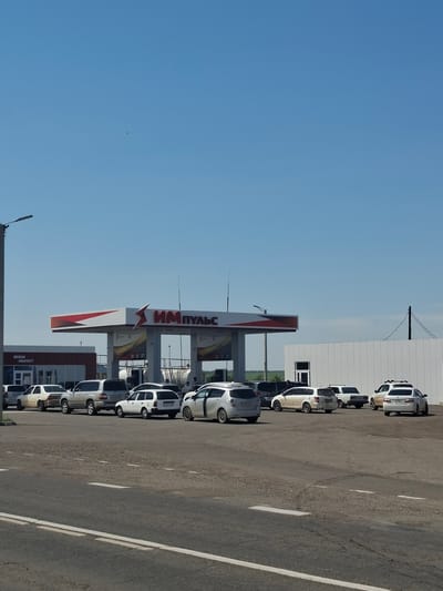 The image shows a gas station with a white and red canopy, labeled "ИМ ПУЛЬС". Several cars are parked in front of the gas pumps and a mini-market building. The gas station appears to be located in Bayanday, Russia, as indicated by the text provided. The sky is clear and blue, and the weather appears to be sunny. The ground is paved with asphalt. The scene depicts a typical gas station environment with vehicles and structures associated with the refueling and convenience services.