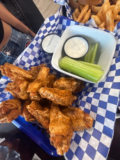 In Sun City Center, United States of America, a close-up overhead shot features a blue plastic basket lined with blue and white checkered paper. Within the basket, a pile of golden-brown chicken wings is seasoned with visible spices. To the right of the wings, a white ceramic bowl holds a creamy dipping sauce and three green celery sticks. In the background, french fries are partially visible. A person's arm, wearing denim shorts and a black top, is visible on the left side of the frame.