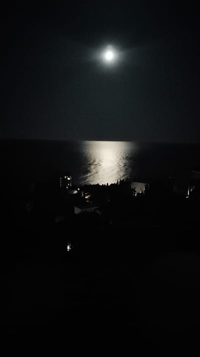 The image depicts a nighttime scene of the sea and a coastal settlement, likely in Кореизский поселковый совет, Ukraine. The full moon is prominently visible in the upper center of the sky, casting a bright reflection on the water. The sea is dark, with the moonlight creating a shimmering path across its surface. In the foreground, the silhouette of a town or village is visible, with scattered lights from buildings indicating human habitation. Trees are also discernible as dark shapes against the slightly lighter sky. The overall atmosphere is serene and tranquil, with the moonlit sea dominating the composition. The image appears to be a photograph taken from an elevated vantage point, offering a panoramic view of the coastline.