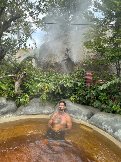 A man with tattoos is sitting in a hot spring, surrounded by lush vegetation. Steam rises from the water and a nearby rock formation. A sign labeled "HERB RO 37-39" is visible amongst the plants. The location is in Ban Khlong Khian, Thailand.