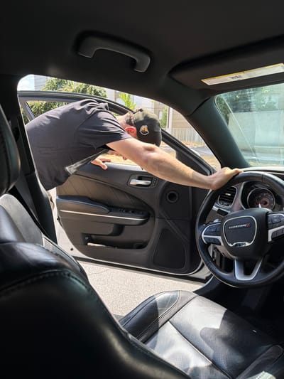 The image shows a man standing next to the open driver's side door of a car. He is wearing a black t-shirt and a baseball cap. His arm is raised and he appears to be interacting with the interior of the car door. The interior of the car, including the steering wheel and part of the seats, is visible. The car appears to be parked outdoors, with sunlight illuminating the scene, suggesting it is daytime. The background shows greenery and a building, consistent with an urban or suburban setting. The image is taken from inside the car, looking outwards, giving a unique perspective. The car has a Dodge emblem on the steering wheel. There is a warning label on the sun visor. The man's posture and focused expression suggest he is engaged in a task related to the car.