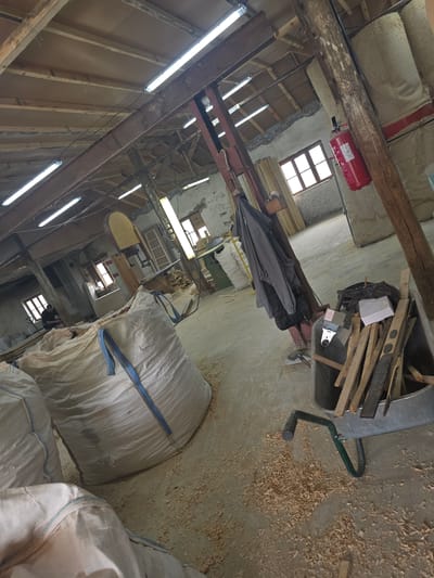 The image depicts a dimly lit, somewhat cluttered interior of what appears to be a workshop or storage area in Kaleytsa, Bulgaria. The scene is dominated by wooden beams, construction materials, and industrial elements. A prominent red fire extinguisher is mounted on a wooden beam near a window with multiple panes.
In the center of the frame, a wheelbarrow is filled with pieces of wood and other debris. Nearby, a dark grey umbrella or tarp is draped over some items. The floor is covered in sawdust, suggesting woodworking or a related activity has taken place. Several large, white, woven bags, possibly containing materials, are scattered around the space, one of them with a blue strap across it. Fluorescent lights are visible along the ceiling, providing the primary illumination.
The overall impression is one of a functional, somewhat untidy workspace. There are no people clearly visible in the image. The time of day is not explicitly indicated, but the artificial lighting suggests it could be indoors at any time. The location cues, such as the style of the building and the potential presence of industrial bags, align with a rural or workshop setting in Bulgaria. No discernible text is clearly legible beyond what's on the fire extinguisher.