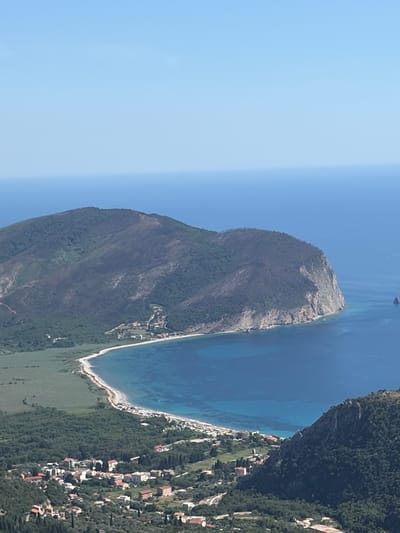 An aerial, high-angle shot captures a coastal scene with a mountainous peninsula jutting into the deep blue Adriatic Sea. The peninsula, covered in dark green vegetation with patches of lighter, possibly burnt, areas, forms a prominent headland. A crescent-shaped beach with white sand and a scattering of beach umbrellas lines the bay, meeting a stretch of green fields and a small town. The town is characterized by numerous buildings with terracotta roofs, nestled amidst dense green trees and some winding roads. The clear blue sky transitions into the slightly lighter blue of the sea. The scene appears to be of the area around Novoselje, Montenegro, with the iconic Sveti Stefan visible in the distance, though not explicitly identified in the image.