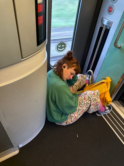The image shows a young person with red hair tied up in a bun, wearing a green sweatshirt and patterned pants, sleeping while leaning against a curved, light-colored wall inside a train. The person is wearing black Vans sneakers and pink socks. A bright yellow bag is placed next to them. The train carriage interior features a green door, a metal pole, and a window showing a blurry green landscape, indicating motion. There are some circular buttons and a smiley face decal on the wall near the window. The floor is carpeted in black. The setting appears to be a train on the route between Loosdorf, Austria, and St. Pölten, Austria, possibly on the Franz-Josefs-Bahn line, given the characteristic interior design and the rural scenery visible through the window.