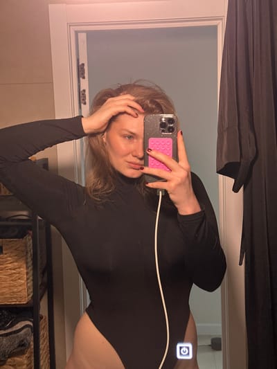 A mirror selfie is captured indoors in the city of Tashevka, Russia. The image features a female individual, depicted from mid-thigh up, with light brown hair and fair skin. She is wearing a form-fitting black long-sleeved turtleneck bodysuit. Her right hand is raised, with fingers touching the top of her head and hair. Her left hand holds a silver iPhone, encased in a glittery clear case, which has a bright pink textured accessory attached to its back. A white charging cable is plugged into the phone's charging port. The subject is looking directly towards the camera/mirror. In the reflected background to the left, a multi-tiered shelving unit with light brown wicker baskets is partially visible. To the right of the subject, a white door frame with dark hinges is present, with a dark, draped garment hanging adjacent to it. A light blue wall is reflected beyond the door frame. The lower portion of the mirror reflects a light-colored floor and a digital power symbol.