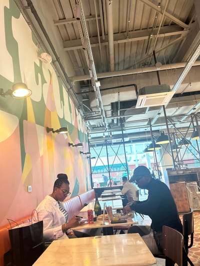 The image captures an indoor scene in Serris, France, featuring a high-ceilinged space with visible industrial infrastructure such as metal conduits, support beams, and ventilation systems. The lower half of the wall is adorned with a vibrant mural composed of geometric shapes and pastel colors. Recessed spotlights are positioned along the wall, and a single, more prominent spotlight is located in the foreground.

In the foreground, a woman wearing glasses and a white striped shirt sits at a table. She is holding a mobile phone in her right hand and appears to be interacting with it. On the table, there are bottles, glasses, and various items suggesting a meal or gathering.

Another individual is partially visible in the background, dressed in black and leaning over the table, seemingly involved in the activities taking place there. The background reveals an open area with additional tables and seating, indicating a restaurant or café setting. The architectural design suggests a blend of industrial and modern elements.