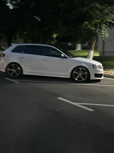 The image displays a white two-door Audi A3 Sportback hatchback parked on an asphalt surface during nighttime in Dzhebel, Bulgaria. The vehicle is oriented with its left side facing the viewer, slightly angled towards the front. It features dark-tinted windows, a subtle roof spoiler, and five-spoke alloy wheels. The front fascia, including the distinctive Audi grille and headlights with integrated LED elements, is visible on the right. The car's ride height appears lowered.
The foreground consists of dark asphalt with white painted lines, indicative of a parking area or road markings. In the background, a strip of illuminated green grass separates the paved area from dense, dark tree foliage. Several tree trunks, appearing light in color, are interspersed within the background. A light-colored building is partially visible on the far right. The scene is illuminated by unseen artificial light sources, creating areas of contrast and shadow. No individuals are present.