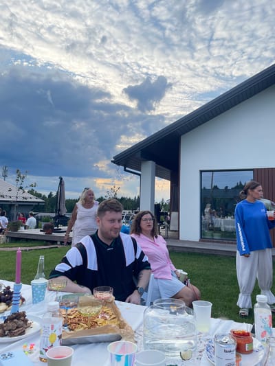 The image depicts an outdoor gathering at a modern house in Simagino, Russia, under a cloudy sky. In the foreground, a white tablecloth is laden with food and drinks, including meat, pretzels, chips, fruit, and various beverages. A man wearing a black and white jacket is seated at the table, facing the camera. To his right, a woman in a pink shirt is also seated. Further to the right, a woman in a blue sweatshirt and white pants is standing and holding a drink. Behind them, other people are visible, some near the house and others further in the background. The house features a white exterior with dark trim and large windows. The surrounding environment includes a well-kept lawn, trees, and a fence. The overall scene suggests a casual social event taking place in a suburban or semi-rural setting.