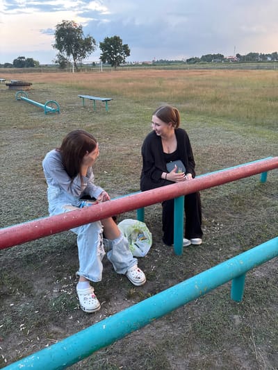 Two young women are seated on a red metal bar in an outdoor recreational area. The woman on the left is wearing ripped light blue jeans and a grey long-sleeved shirt. She has her hand over her face and appears to be laughing or crying. The woman on the right is wearing black wide-leg pants and a black open-front jacket over a dark top. She is holding a small dark clutch and smiling. A plastic bag is on the ground next to her. The background shows a field with dry grass, trees, and a distant village with buildings and a church spire. The sky is overcast with some hints of sunset colors. The overall impression is a casual, outdoor gathering in a rural setting. This location appears to be in or around the village of Sarotski, within the Sarotski Sel'ski Sovet in Belarus.