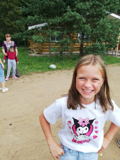 In the foreground, a young girl with long brown hair and blue eyes smiles at the camera. She is wearing a white t-shirt with a graphic of Kuromi, a Sanrio character, and light blue denim shorts. Glitter is applied to her cheeks. In the background, an older woman in a purple and white patterned shirt stands with her hands on the shoulders of a young girl wearing a red long-sleeved shirt and gray pants. To the right of them is a wooden cabin, and pine trees and greenery surround the area. The ground is covered in sand and sparse grass. The scene appears to be outdoors, possibly in a rural or recreational area near the city of Shapki, Russia.