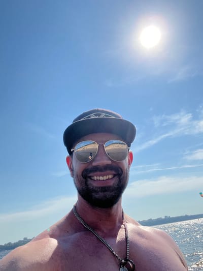 A man wearing a hat and sunglasses is taking a selfie outdoors. The sun is bright in the sky, and there is water in the background. The man has a beard and is smiling. He is wearing a necklace. The reflection in his sunglasses shows a beach scene with people. The location is not specified in the image or in the provided context for the city of Sosnovka, Russia.