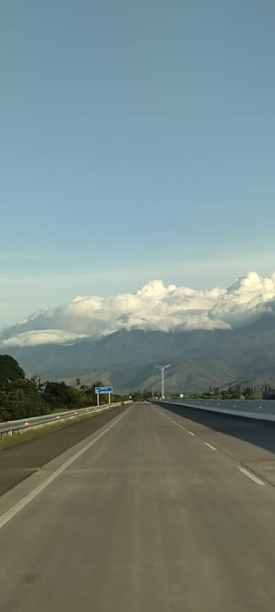 An overcast sky stretches above a highway in Baksukon, Indonesia, with a mountain range visible in the distance. The highway, with its clear lane markings and concrete barriers, stretches into the horizon, suggesting a long and open road. Trees and vegetation line the sides of the road, with some taller trees reaching towards the sky. The overall scene is serene and suggests a journey through a natural landscape.