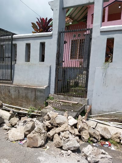 This image depicts the exterior of a residential property in Viengthong, Laos, viewed from a slightly elevated ground level. The foreground is dominated by a substantial pile of light grey concrete rubble and stones scattered across an asphalt surface, with small pieces of litter, including wrappers, visible among the debris. Beneath and adjacent to the rubble, several white plastic pipes are exposed.

The midground features a light grey, rendered concrete perimeter fence. This fence incorporates three rectangular vertical openings on its left side and a lower concrete ledge. A dark-colored, vertical-bar metal gate, appearing slightly ajar, is centrally positioned, leading to an elevated concrete step area overgrown with sparse vegetation. To the left of the main gate, a narrower, similar vertical-bar metal section suggests an additional gate or fence panel. A wooden plank rests on the concrete step near the main gate.

In the background, behind the fence and gate, a section of a light pink building, presumed to be a house, is visible. It features a brown-framed window with multiple panes. A sloped roof structure with green and terracotta-colored tiles is discernible above the house section. A large plant with reddish-purple foliage is prominently situated behind the left side of the concrete fence, extending above its top edge. The sky above is overcast. No individuals are present in the scene, and no overt actions or interactions are depicted.