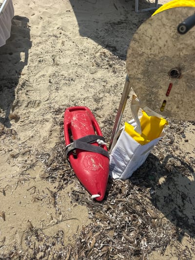 A red rescue buoy lies on a sandy beach in Castiadas, Italy, with a white bag containing yellow flotation devices and a portion of a metal umbrella pole nearby. The scene is illuminated by sunlight, casting shadows on the sand. The beach is littered with dried seaweed and other natural debris. The buoy is positioned at an angle, and its black strap is visible. The bag is open, revealing its contents. The umbrella pole is upright, with a circular metal disc at its top, partially obscuring the sky.